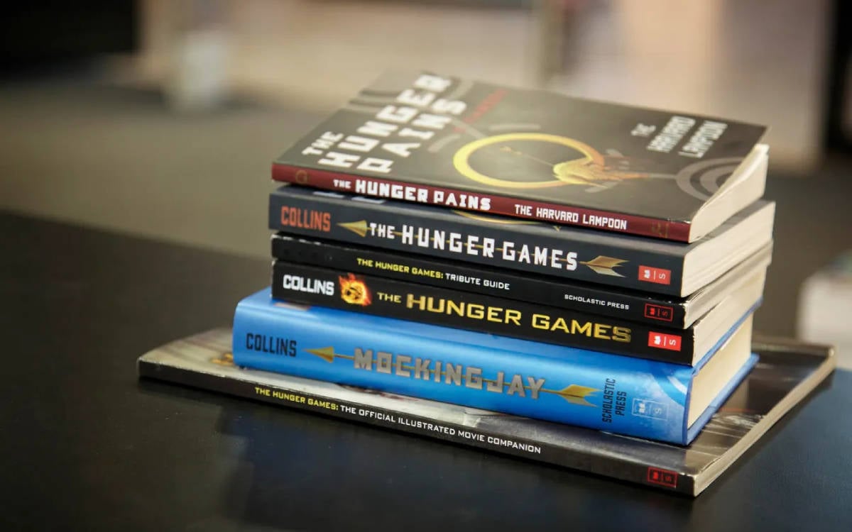 Görselde, bir masanın üzerine üst üste dizilmiş birkaç kitap bulunuyor. Bu kitaplar arasında "The Hunger Games" (Açlık Oyunları) serisine ait kitaplar yer alıyor. En üstte, "The Hunger Pains" adlı bir parodi kitabı var. Hemen altında ise Suzanne Collins'in yazdığı ünlü "The Hunger Games" kitapları bulunuyor: "The Hunger Games", "The Hunger Games: Tribute Guide" ve "Mockingjay". En altta ise "The Hunger Games: The Official Illustrated Movie Companion" kitabı yer alıyor. Kitapların yan yana dizilişi, serinin popülerliğini ve çeşitliliğini gösteriyor.