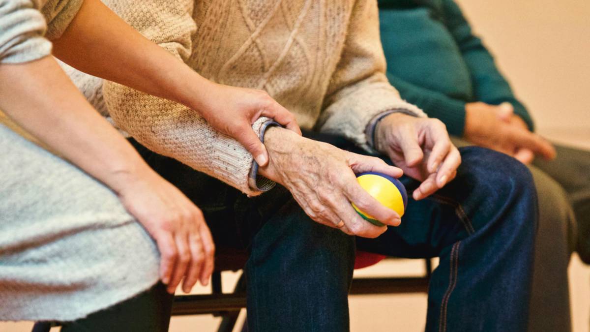 Görselde, bir yaşlı kişinin elleri odak noktasıdır. Yaşlı kişi dizlerinde oturmakta ve elinde sarı-mavi bir top tutuyor. Kişinin üzerinde krem rengi örgü desenli bir kazak bulunuyor. Yanında ise başka bir kişi, yaşlı kişinin koluna nazikçe dokunarak ona destek veriyor. Bu ikinci kişinin sadece elleri ve kolları görünüyor, gri renkte sade bir kıyafet giymiş. Yaşlı kişinin yanında bir başka kişi daha oturuyor, ancak bu kişinin sadece kolları ve kısmen üst bedeni görünüyor, yeşil bir kazak giymiş. Görüntü, bir bakım ortamını ya da destekleyici bir durumu yansıtıyor; sıcak ve şefkat dolu bir anın fotoğrafı olarak öne çıkıyor.