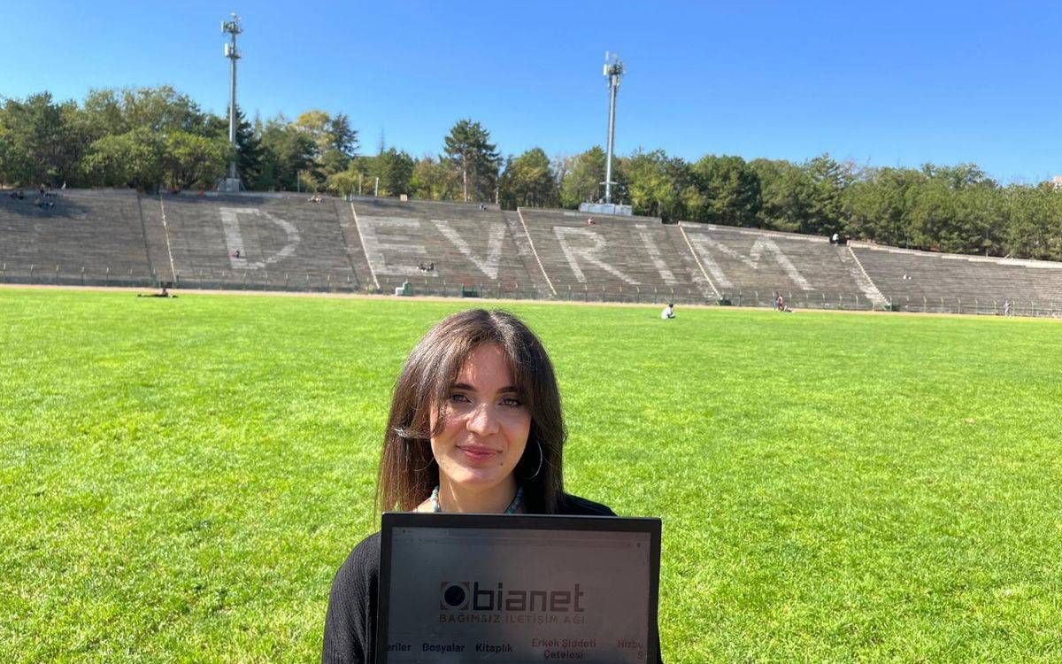 Fotoğrafta, geniş bir yeşil alanın önünde bir kadın duruyor. Kadın, elinde "Bianet Bağımsız İletişim Ağı" yazılı bir tablet tutuyor. Arka planda büyük bir stadyum tribünü görülüyor ve tribün sıralarının üzerine büyük harflerle "DEVRİM" yazılmış. Hava açık ve güneşli, gökyüzü masmavi. Tribünlerin arkasında ağaçlar yer alıyor, ve stadyumda birkaç kişi uzakta oturuyor. Kadının saçları açık ve hafif gülümseyen bir ifadesi var. Fotoğraf, açık bir alanda güzel bir sonbahar gününü yansıtıyor.