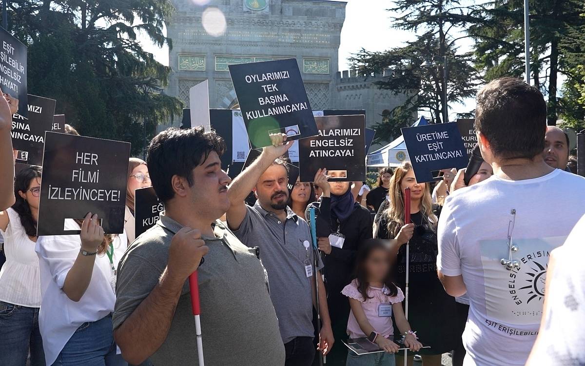 Görselde, engelli bireylerin haklarını savunmaya yönelik bir eylem yapıyor. Bir grup insan ellerinde pankartlarla bir protestoediyor. Katılımcılar arasında çeşitli yaş gruplarından insanlar yer alıyor. Bazı pankartlarda "Her filmi izleyinceye!", "Oylamalarımızı engelsiz atıncaya!" ve "Üretime eşit katılıncaya!" gibi ifadeler yer alıyor.