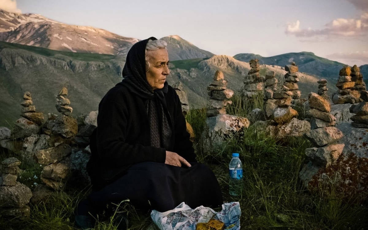 Fotoğrafta, dağlık bir alanda yaşlı bir kadın görülüyor. Kadın, başına siyah bir örtü takmış, koyu renkli kıyafetler giymiş ve yere oturmuş durumda. Önünde bir gazete kağıdına sarılı yiyecekler ve bir su şişesi bulunuyor. Etrafında ise üst üste dizilmiş küçük taş yığınları var; bu taş yığınları, dağlık bölgelerde yapılan sembolik düzenlemeleri anımsatıyor.  Arka planda ise yüksek dağlar ve doğa manzarası görülüyor. Dağların zirvesinde kar parçaları mevcut, manzara oldukça sakin ve huzurlu bir atmosfer yayıyor. Fotoğraf, gün batımında veya sabah erken saatlerde çekilmiş gibi, çünkü ışık yumuşak ve alacakaranlık hissi veriyor. Kadının bakışları düşünceli ve huzurlu, doğayla iç içe bir ortamda derin düşüncelere dalmış gibi görünüyor.