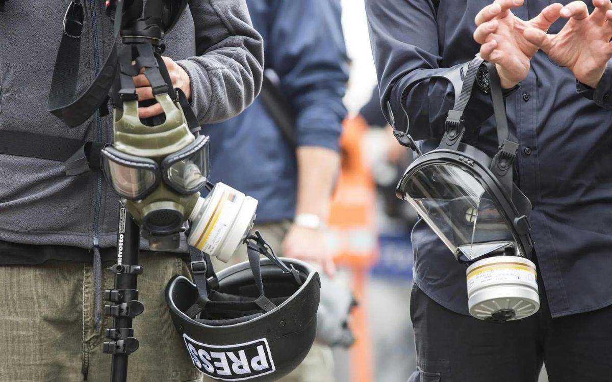 Bu fotoğrafta, ellerinde koruyucu gaz maskeleri tutan ve gazetecilikle ilgilenen kişilerin görüntüsü yer almakta. Ön planda bir kişinin elinde siyah bir kask var, kaskın üzerinde "PRESS" (basın) yazısı dikkat çekiyor. Diğer kişiler ise gaz maskeleri ve gaz filtreleri taşımakta. Fotoğrafın genel atmosferi bir protesto ya da riskli bir alanda çalışan gazetecileri yansıtıyor olabilir. Kıyafetler ve ekipmanlar, kişilerin zorlu koşullara karşı korunma amacıyla hazırlandığını gösteriyor.