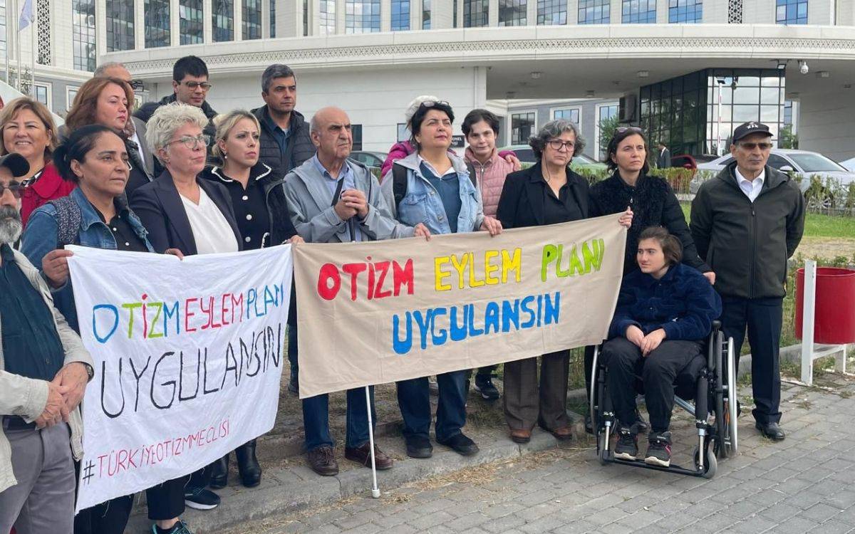 Bu fotoğraf, bir grup insanın toplu halde bir araya geldiği bir protesto veya farkındalık etkinliğini gösteriyor. Grup, ellerinde pankartlar tutarak bir arada duruyor. Pankartlarda büyük harflerle "OTİZM EYLEM PLANI UYGULANSIN" yazıyor. Katılımcıların çoğu ayakta dururken, ön planda tekerlekli sandalyede oturan bir genç bulunuyor. Grup farklı yaşlardan ve cinsiyetlerden bireylerden oluşuyor, arka planda ise büyük ve modern bir bina görünüyor.  Bu etkinlik, otizm eylem planının uygulanması talebiyle yapılan bir gösteri gibi görünüyor ve sosyal haklar veya sağlık politikalarıyla ilgili bir talebi dile getiriyor. Pankartlarda yazan ifadeler, bu taleplerin açık bir şekilde ifade edildiğini gösteriyor.