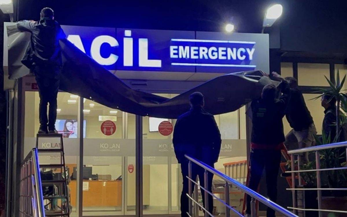 Görselde bir hastanenin "Acil" (Emergency) girişine ait tabela görülüyor. "Acil" ve "Emergency" yazıları mavi ve beyaz renklerle ışıklandırılmış durumda. Görselin ön planında, birkaç kişi büyük bir örtü ya da pankart gibi bir malzemeyi tutarak tabelayı kapatmaya çalışıyor.