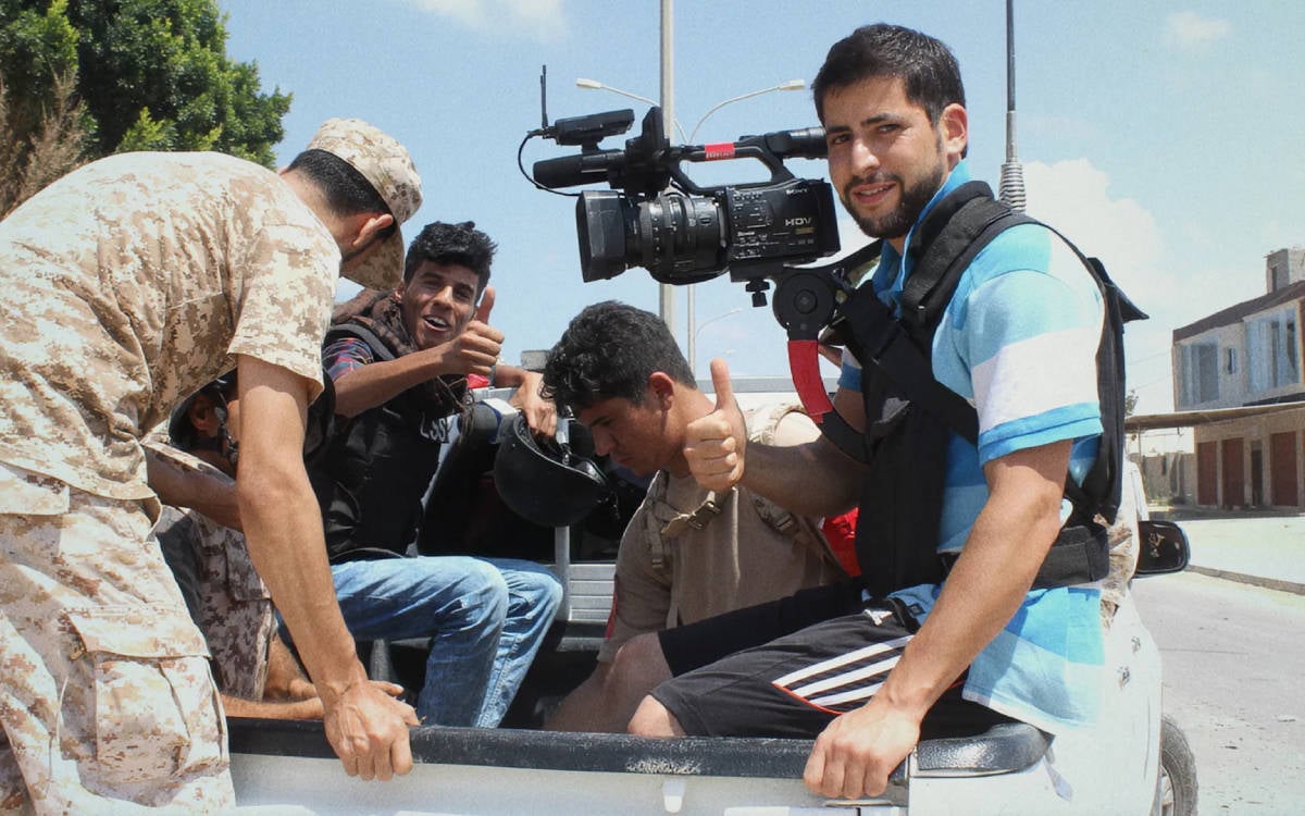 Fotoğrafta, açık bir aracın kasasında seyahat eden bir grup insan görülüyor. Arka planda gökyüzü açık ve hava güneşli. Önde, mavi-beyaz çizgili bir tişört giyen ve omzunda büyük bir video kamera taşıyan bir gazeteci veya kameraman var. Kameraman doğrudan kameraya bakarak hafifçe gülümsüyor ve başparmağını kaldırarak olumlu bir işaret yapıyor.  Aracın kasasında ayrıca kamuflaj desenli kıyafetler giymiş bir asker ve sivil giyimli birkaç kişi daha bulunuyor. Bu kişiler de kameraya başparmaklarıyla "tamam" işareti yaparak poz veriyorlar. Ortam, dostane ve rahat bir havada; grup arasında gerginlik değil, aksine rahat bir iletişim havası hakim. Görüntü, çatışma bölgelerinde görev yapan gazetecilerin çalışmalarına dair bir anı yansıtıyor.