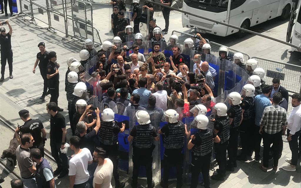 Fotoğrafta, çevik kuvvet polisleri ve bir grup protestocu veya gazeteci arasında bir çatışma veya yoğun bir tartışma anı görülüyor. Polisler, kalabalığı çevreleyerek ellerinde taşıdıkları şeffaf kalkanlarla bir bariyer oluşturmuş durumda. Polisler kask takmış ve sırtlarında "POLİS" yazılı üniformalar var. Kalabalık, polis çemberinin içinde yer alıyor ve bazıları ellerinde cep telefonları veya kameralarla görüntü almaya çalışıyor.  Bu olay, bir protesto, basın açıklaması veya toplumsal bir meseleye dair yapılan bir eylem sırasında yaşanmış gibi görünüyor. Polisler, kalabalığı kontrol altında tutmaya çalışırken, çevrede bazı izleyiciler ve diğer polis memurları da bulunuyor. Çevredeki bariyerler ve polislerin düzenli dizilimi, güvenlik önlemlerinin sıkı bir şekilde alındığını gösteriyor.