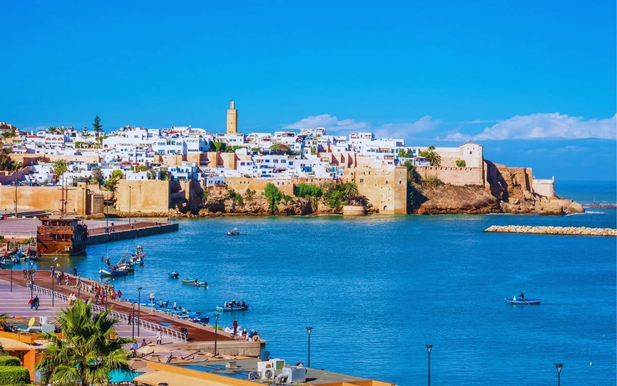 Fotoğrafta, Fas’ın başkenti Rabat'taki sahil bölgesi görülüyor. Denize nazır yerleşim yerleri ve tarihi surlarla çevrili eski şehir, beyaz ve pastel tonlarda sıralanmış evlerden oluşuyor. Bu evlerin arasında bir minare veya kule de göze çarpıyor, bu da muhtemelen eski bir cami veya başka bir tarihi yapının parçası.  Ön planda, su üzerinde balıkçı tekneleri veya küçük sandallar yer alıyor. Sahilde insanlar yürüyüş yapıyor, manzaranın tadını çıkarıyor. Deniz parlak mavi bir renkte, ve genel atmosfer canlı ve sıcak bir tatil beldesini andırıyor. Gökyüzü berrak ve masmavi, birkaç küçük bulutla birlikte açık bir hava hakim.  Bu tür bir manzara, tarihi ve kültürel zenginliklerle modern yaşamın iç içe geçtiği, sahil kıyısında huzurlu bir yaşamı yansıtıyor.