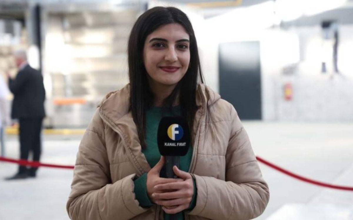 u fotoğrafta, bir kadın gazeteci görülüyor. Kadın, açık renkli bir mont giymiş ve yeşil bir üst giysiyle kombinlemiş. Elinde, üzerinde "Kanal Fırat" logosu bulunan bir mikrofon tutuyor. Saçları düz ve omuz hizasında. Kadın, objektife bakarak hafif bir gülümseme sergiliyor. Arka planda flulaştırılmış bir iç mekan görülüyor; bu ortamda bir kırmızı kurdele ve birkaç insan figürü yer alıyor, ancak detaylar net değil. Fotoğraf, muhtemelen bir haber ya da röportaj sırasında çekilmiş.