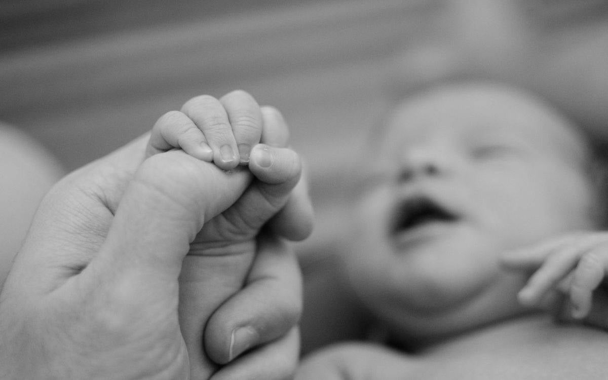 Bu siyah-beyaz fotoğraf, bir yetişkinin elini nazikçe kavrayan yenidoğan bir bebeğin minik elini gösteriyor. Bebek net bir şekilde görünmese de, arka planda flu şekilde yatan bir bebek olduğu anlaşılıyor. Görsel, yeni doğmuş bir bebeğin hassasiyetini ve korunmaya muhtaçlığını sembolize ederken, aynı zamanda dokunaklı bir bağ kurma anını yansıtıyor. Fotoğrafın siyah-beyaz oluşu, duygusal etkisini güçlendiriyor ve zamansız bir estetik katıyor.