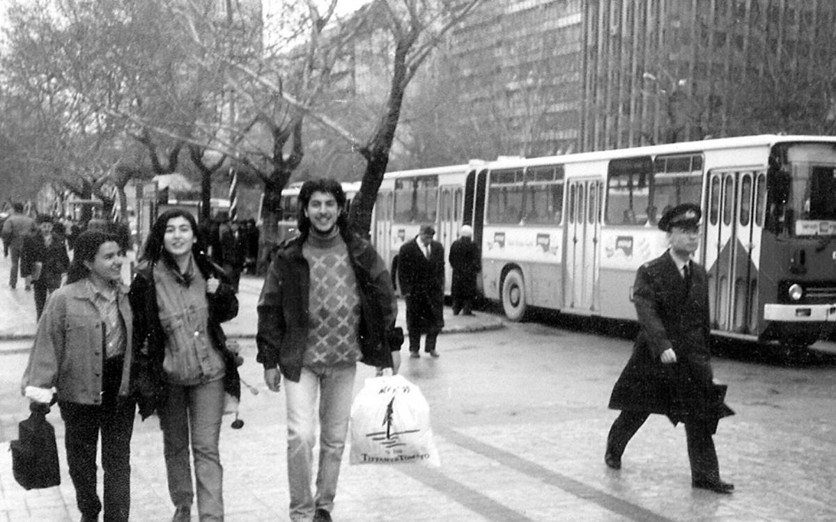 Fotoğrafın ön planında üç kişi birlikte yürüyor. Soldaki kişi kısa saçlı, üzerinde açık renkli bir ceket var ve elinde bir çanta taşıyor. Ortadaki kişi uzun saçlı, gülümseyerek kameraya bakıyor; üzerinde düğmeli bir gömlek ve açık renk bir ceket bulunuyor. Sağdaki kişi ise genç bir adam, dalgalı saçları var, kazak ve kalın bir ceket giymiş, elinde bir plastik poşet taşıyor. Poşetin üzerinde bir ağaç silueti ve yazılar yer alıyor.  Arka planda bir şehir manzarası görülüyor. Sol tarafta kaldırım boyunca dizilmiş ağaçlar yer alırken, sağda ise park etmiş veya hareket halinde olan eski model otobüsler var. Birkaç kişi otobüslere binmek veya inmek için bekliyor gibi görünüyor. Sağ köşede, bir polis memuruna benzeyen, üniforma giymiş bir kişi yürümekte.