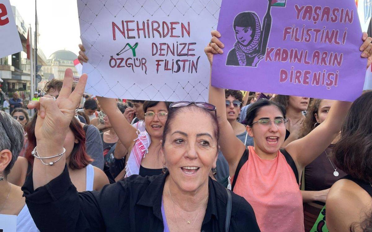 Bu fotoğrafta, Filistin’e destek amaçlı düzenlenen bir protesto gösterisi görülüyor. Ön planda siyah kıyafetli bir kadın, gülümseyerek zafer işareti yapıyor. Yüzünde kararlılık ve coşku ifadesi var.  Hemen arkasında, iki kadın dikkat çekiyor. Sol taraftaki kadın beyaz bir pankart tutuyor. Pankartta kırmızı ve yeşil harflerle “Nehirden Denize Özgür Filistin” yazılı. Sağ tarafta ise başka bir kadın mor renkli bir pankart kaldırmış; üzerinde başörtülü, elinde bayrak tutan bir kadın figürü ve “Yaşasın Filistinli Kadınların Direnişi” yazısı yer alıyor. Her iki pankart da Filistin’e destek mesajı taşıyor.