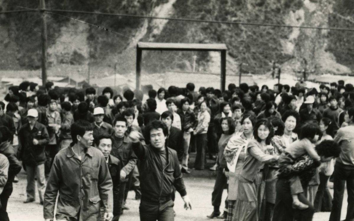 Fotoğrafta büyük bir kalabalık açık bir alanda toplanmış. Görüntü siyah-beyaz ve tarihî bir dönemden olduğu izlenimi veriyor. Ön planda bir grup insan dikkat çekiyor; bazıları ellerini havaya kaldırmış ya da bir yönü işaret ediyor. Kalabalıkta kadınlar, erkekler ve çocuklar var. Bir kadın, küçük bir çocuğu kucağında taşıyor. Arkada dağlık bir alan ve çadır benzeri yapılar seçiliyor. İnsanların yüz ifadeleri genel olarak ciddi ve dikkatli görünüyor. Fotoğraf, bir protesto, yürüyüş veya kitlesel bir hareket sırasında çekilmiş gibi duruyor ve dayanışma ya da mücadele teması yansıtıyor.