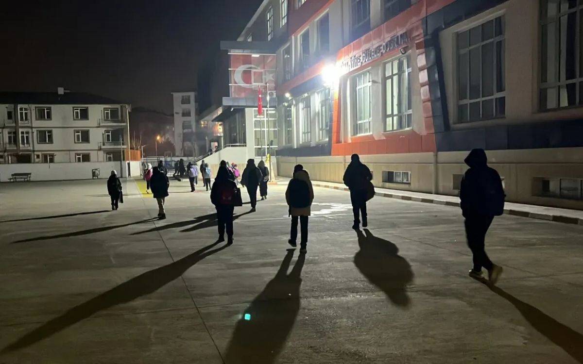 Bu fotoğrafta, bir grup insan, gece karanlığında bir okulun bahçesinde yürümekte. Ortam oldukça loş ve okulun üzerindeki lambalar mekânı aydınlatıyor. Yerde yürüyen kişilerin uzun gölgeleri belirgin şekilde görünüyor. İnsanlar sırt çantaları taşıyor ve çoğunlukla kışlık kıyafetler giymiş. Okul binası modern bir yapıya benziyor ve binanın üzerinde Türk bayrağı ile birlikte "Cumhuriyet" kelimesinin yazdığı bir tabela dikkat çekiyor. Arka planda apartmanlar ve sakin bir şehir atmosferi görülmekte. Fotoğraf, erken saatlerde ya da geç bir saatte çekilmiş izlenimi veriyor.