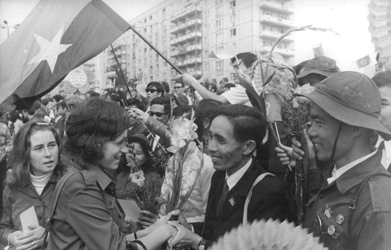 Bu siyah-beyaz fotoğraf, büyük bir kalabalığın içinde gerçekleşen coşkulu bir karşılamayı veya kutlamayı gösteriyor. Ön planda, genç bir kadın ve takım elbiseli bir adam el sıkışırken gülümsüyorlar. Kadının elinde çiçekler var ve adamın yakasında küçük bir rozet dikkat çekiyor.  Yanlarında, askeri kıyafet giymiş ve Vietnam Savaşı döneminde Kuzey Vietnam ordusunun kullandığı tipte bir kask takan bir adam bulunuyor.