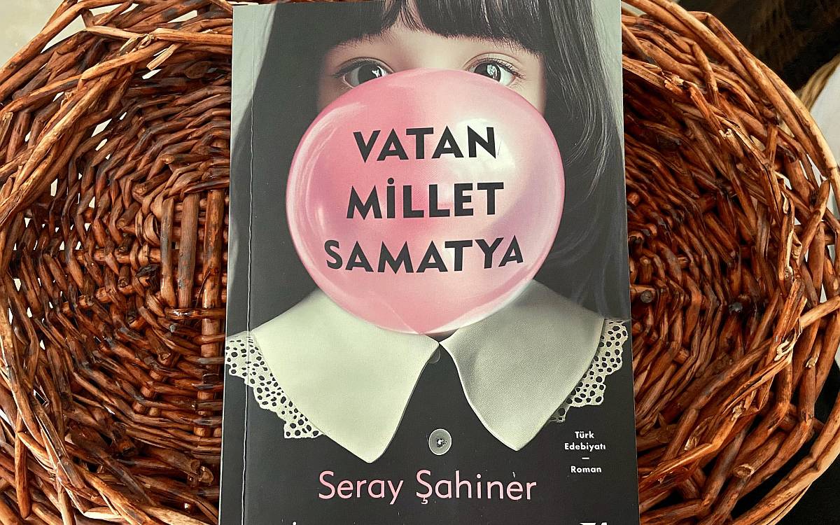 Görselde Seray Şahiner’in Vatan Millet Samatya adlı kitabı yer alıyor. Kitabın kapağında, siyah saçlı bir kız çocuğunun yüzü yer alıyor. Kız, büyük ve pembe bir balon sakızı şişirmiş ve bu sakızın üzerine kitabın adı, "Vatan Millet Samatya" yazıyor. Sakız, çocuğun ağzını ve burun bölgesini kapatırken, sadece gözleri açık görünüyor.