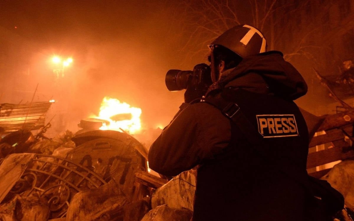 Fotoğraf, kaotik ve tehlikeli bir ortamda çalışan bir basın mensubunu gösteriyor. Ön planda, sırtında "PRESS" (Basın) yazılı bir yelek giyen bir gazeteci yer alıyor. Başında koruyucu bir miğfer var. Kalın bir mont giymiş ve elinde profesyonel bir fotoğraf makinesiyle olayları belgelemeye çalışıyor. Arka planda, büyük bir yangın var; alevler ve yoğun duman gökyüzünü kaplamış. Yanan bir araç ve etrafa dağılmış metal ve beton parçaları görülüyor. Ortamda büyük bir yıkım var, çevrede devrilmiş bariyerler, hurda metal parçaları ve molozlar dikkat çekiyor.