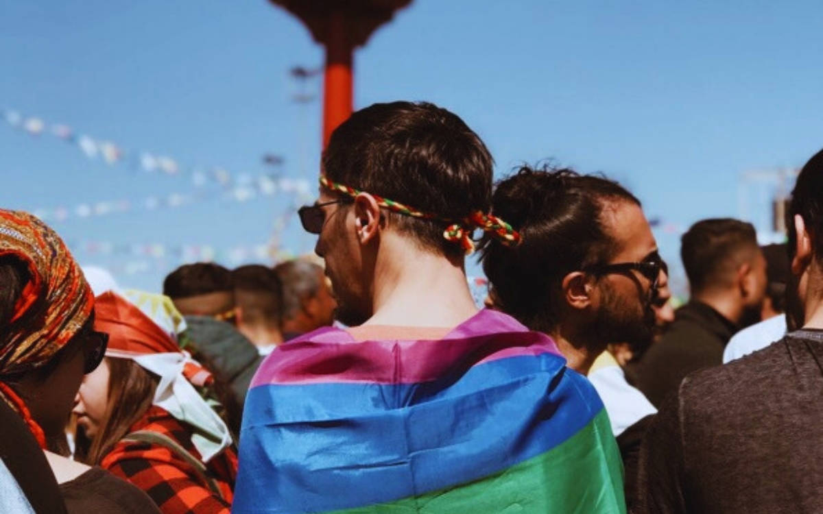 Fotoğrafta 2019 Diyarbakır Newrozu'ndan bir kare var. Ön planda, sırtı kameraya dönük bir kişi LGBTİ+ bayrağı omzuna sarmış. Başında kırmızı, sarı ve yeşil renklerde örgülü bir bant var ve siyah güneş gözlüğü takıyor.