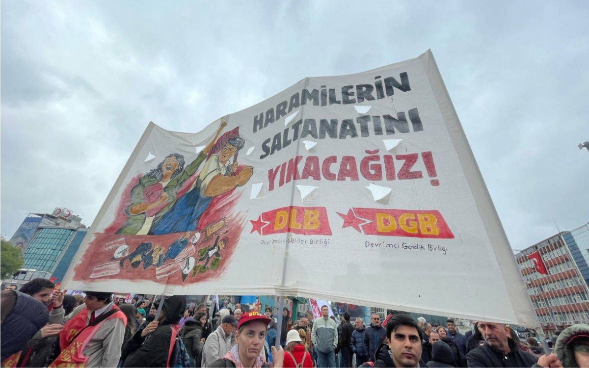 Fotoğrafta, kalabalık bir 1 Mayıs mitinginde çekilmiş bir kare yer alıyor. Görselin merkezinde büyükçe bir pankart dikkat çekiyor.