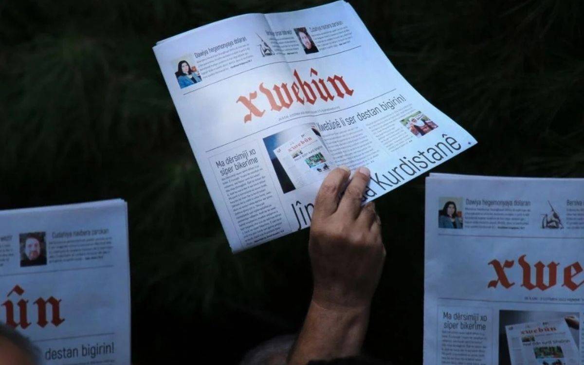 Bu fotoğraf, birkaç kişinin Kürtçe bir gazete okuduğu anı belgeliyor. Görselin odak noktasında, "Xwebûn" adlı gazete bulunuyor. Gazetenin adı, kırmızı renkle gotik stile yakın bir yazı karakteriyle manşet kısmında dikkat çekici biçimde yer alıyor. Başlıklar ve içerikler Kürtçe yazılmış.
