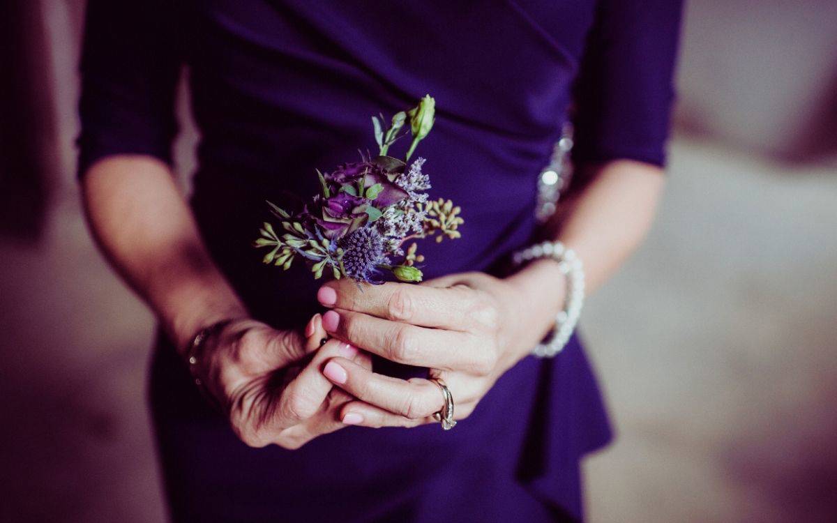 Bu fotoğraf, estetik açıdan zarif ve duygusal bir anı yansıtan yakın plan bir görüntüdür. Koyu mor (veya lacivert) şık bir elbise giymiş bir kadın, iki eliyle küçük bir çiçek demetini nazikçe tutmaktadır. Çiçek demeti mor ve lavanta tonlarında çiçeklerden oluşmuştur; aralarında belki de okaliptüs yaprakları ve minik süs bitkileri de yer alıyor. Çiçeklerin rengi, kadının elbisesiyle uyum içindedir.