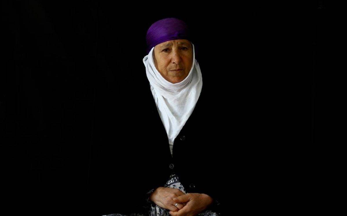 Bu fotoğraf, sade ama etkileyici bir portre çalışmasıdır. Siyah arka plan önünde oturan yaşlı bir kadın yer alır. Kadın geleneksel kıyafetler giymektedir. Başında mor bir örtü, onun altında beyaz bir başörtüsü vardır. Üzerinde siyah düğmeli bir ceket ya da hırka bulunmakta, alt kısmında ise desenli bir etek görünmektedir.