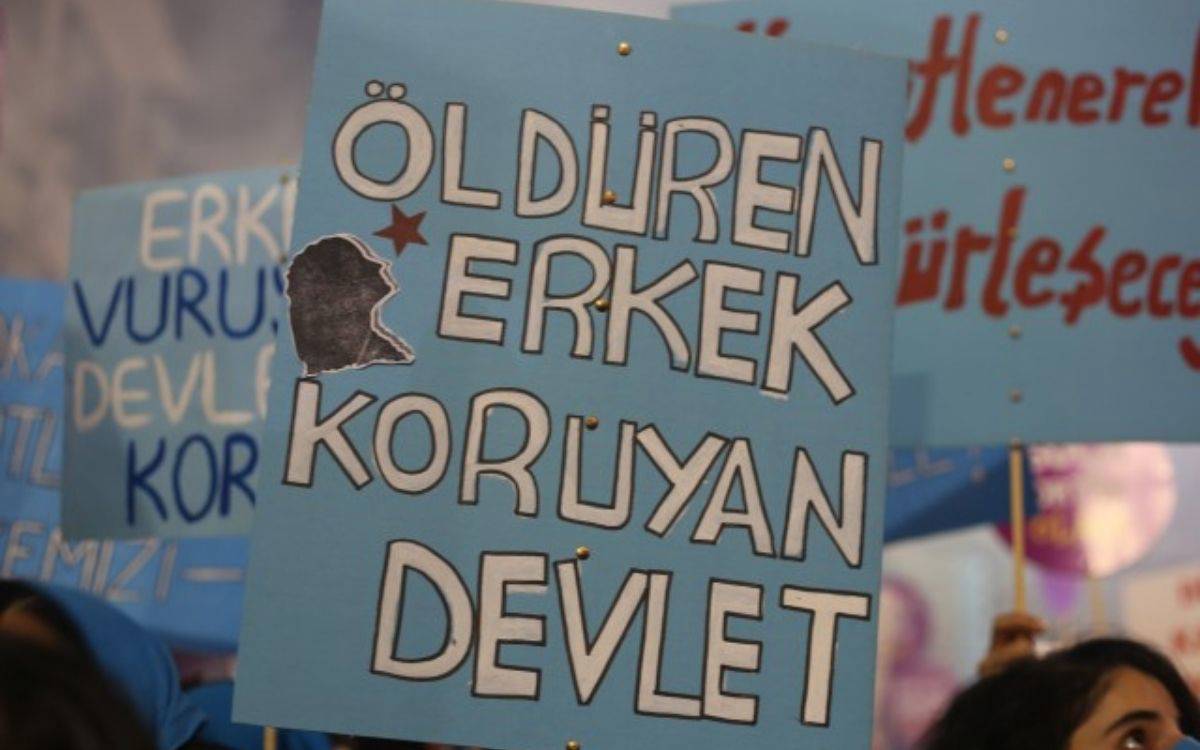 Bu görselde, bir protesto veya eylem sırasında taşınan bir pankart yer alıyor. Pankart açık mavi bir zemin üzerine beyaz ve siyah harflerle yazılmış. Üzerinde şu ifadeler bulunuyor:  "Öldüren erkek, koruyan devlet"