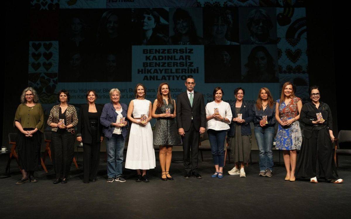 u fotoğraf, 28. Uluslararası Kadın Filmleri Festivali kapsamında düzenlenen özel bir etkinlikten, ödül ya da anı takdimi anını yansıtıyor. Sahne üzerinde yan yana dizilmiş 12 kadın ve 1 erkek yer alıyor. Arka fonda büyük bir ekranda festivalin bu yılki sloganı açıkça görülüyor: “Benzersiz Kadınlar Benzersiz Hikâyelerini Anlatıyor”  Detaylı Betimleme: Kadınlar farklı yaşlarda ve stillerde, kendi doğallıklarıyla sahnede yer alıyor. Kimisi siyah elbise, kimisi ceket-pantolon, kimisi de yazlık bir beyaz ya da desenli elbise giymiş.  Erkek katılımcı takım elbise ve kravatla sahnede, ortada duruyor.  Kadınların neredeyse tamamı ellerinde ödül ya da plaket benzeri objeler tutuyor; bu da etkinliğin bir ödül veya onurlandırma töreni olduğunu düşündürüyor.  Arka ekranda kadınların siyah beyaz portreleri, festival görselleri ve desenlerle bezenmiş bir tasarım yer almakta.  Sahnedeki enerji, sıcaklık ve dayanışma hissi fotoğrafa yansıyor. Kadınların duruşları özgüvenli, gururlu ve içten bir ifadey