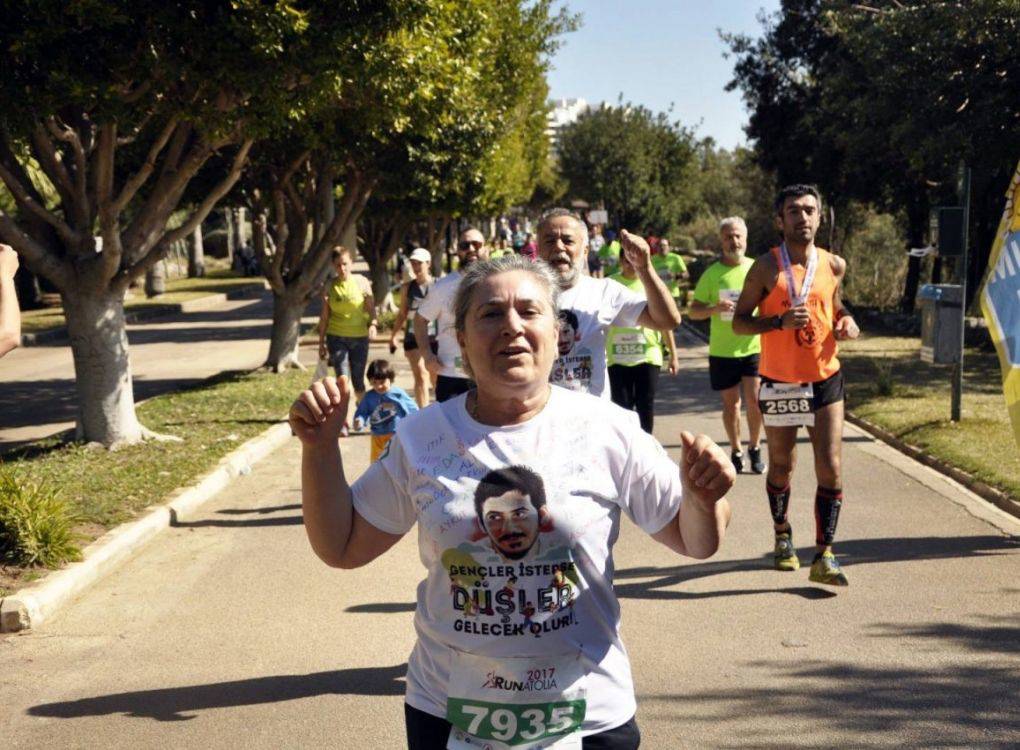 Bu görsel, açık havada bir koşu etkinliğine katılan bir grup insanı gösteriyor. Ön planda, beyaz bir tişört giymiş yaşlıca bir kadın koşuyor. Tişörtünde "Gençler İsterse, Düşler Geleceğe Uçar" yazılı ve bir futbolcunun resmi bulunuyor. Kadın, koşuya katılan diğer insanlara göre daha yavaş bir hızda ilerliyor ve arka planda daha genç koşucular yer alıyor.