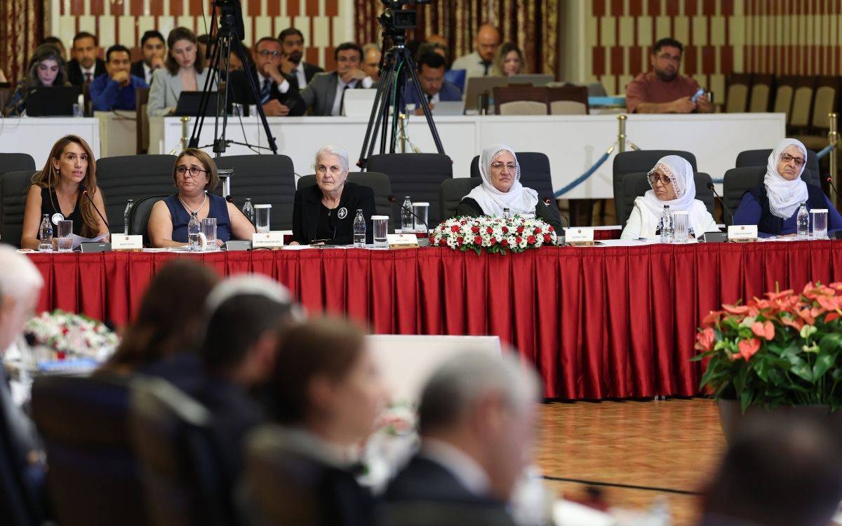 Fotoğrafta resmi bir toplantı salonunda uzun bir masanın arkasında oturan kadınlar görülüyor Masanın üzeri kırmızı örtüyle kaplı ve üzerinde isimlikler su şişeleri mikrofonlar ve çiçek süslemeleri yer alıyor Kadınlardan bazıları başörtülü bazıları başı açık ve hepsi ciddi bir şekilde toplantıya odaklanmış görünüyor Soldaki kadın elinde kağıt tutarak konuşma yapıyor gibi görünüyor diğerleri ise dikkatle dinliyor