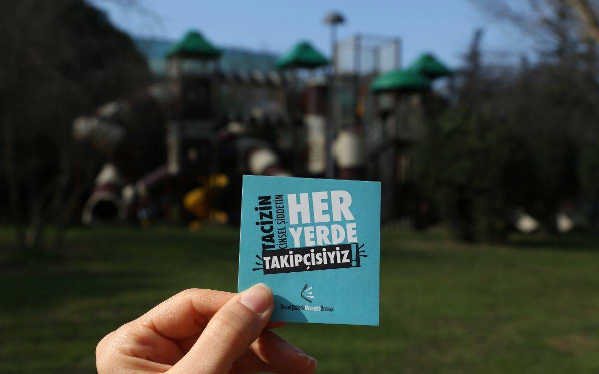 Fotoğrafta bir el, açık mavi renkte bir karton tutuyor. Kartonun üzerinde şu yazı bulunuyor: "Tacizin (cinsel şiddetin) her yerde takipçisiyiz!" Alt kısmında ise küçük puntolarla "Cinsel Şiddetle Mücadele Derneği" ifadesi yer alıyor. Arka planda, bulanık şekilde görünen bir çocuk parkı var. Parkta kaydıraklar ve tırmanma alanları dikkat çekiyor. Çim alanın ve ağaçların bulunduğu park, gündüz vakti, açık havada çekilmiş gibi görünüyor.