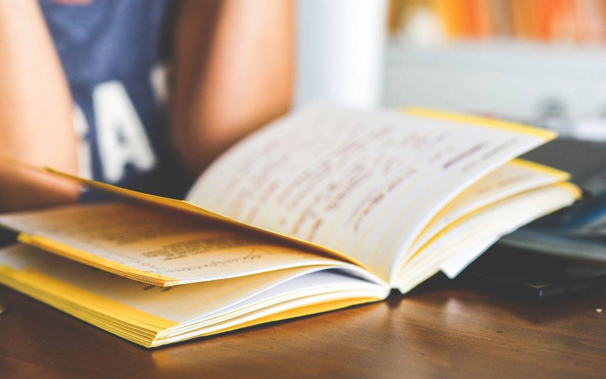 Fotoğrafta bir masa üzerinde açık duran bir defter görülüyor. Defterin sayfaları sarı kenarlıklı ve içinde elle yazılmış notlar veya ders çalışması olduğu anlaşılıyor. Yazılar mavi ve mor tonlarında kalemle yazılmış. (Bu betimleme yapay zeka ile yapıldı)