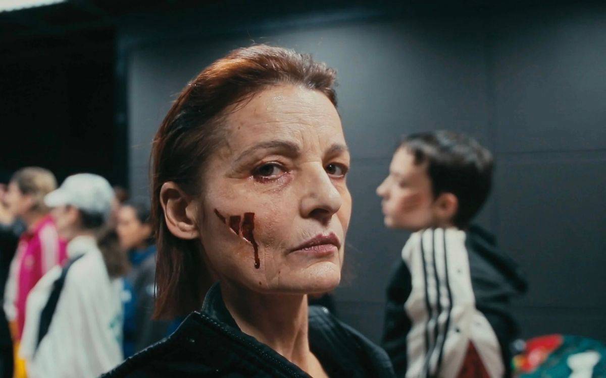 Bir kadın yüzünün üzerinde Adidas'ın sembolü üç çizgi var. Kadın siyah eşofman giyinmiş, kısa saçlı ve kumral.