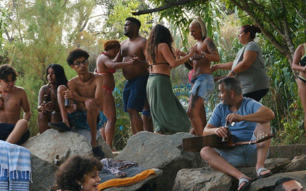 Görselde doğa içinde, kayalık bir alanda toplanmış bir grup insan görülüyor. Ortam rahat, samimi ve yaz mevsimini çağrıştırıyor. Çoğu kişi mayo veya hafif yazlık kıyafetler giymiş. Bazıları kayaların üzerinde oturmuş, ellerinde içecek şişeleri var; diğerleri ayakta dans ediyor veya sohbet ediyor.