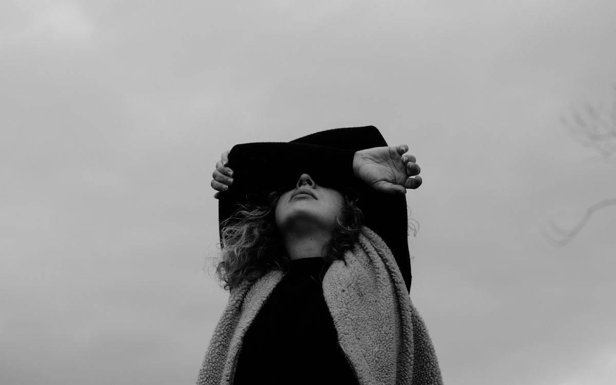 Fotoğraf siyah-beyaz tonlarda çekilmiş. Kadrajda, gökyüzünün açık gri tonları ön planda; bulutlu ve melankolik bir atmosfer yaratıyor. Görselde bir kişi, başını hafifçe yukarı kaldırmış, elleriyle geniş kenarlı şapkasını tutuyor. Yüzünün bir kısmı gölgede kalmış, bu da fotoğrafa gizemli bir hava katıyor.