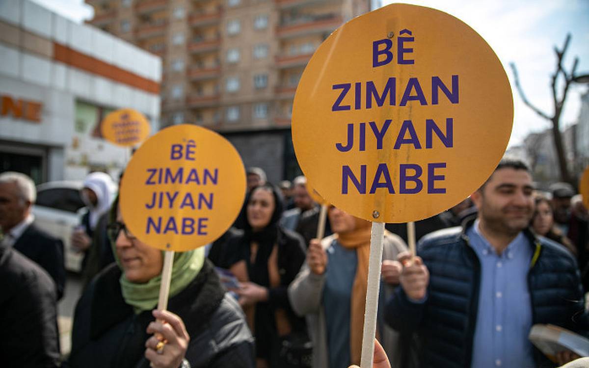 Fotoğrafta bir grup insanın katıldığı açık hava eylemi görülüyor. Kalabalıktaki kişiler ellerinde turuncu zemin üzerine mor harflerle yazılmış yuvarlak dövizler taşıyor. Dövizlerde Kürtçe “Bê ziman jiyan nabe” ifadesi yer alıyor; bu söz Türkçeye “Dilsiz yaşam olmaz” ya da “Dil olmadan hayat olmaz” şeklinde çevrilebilir.