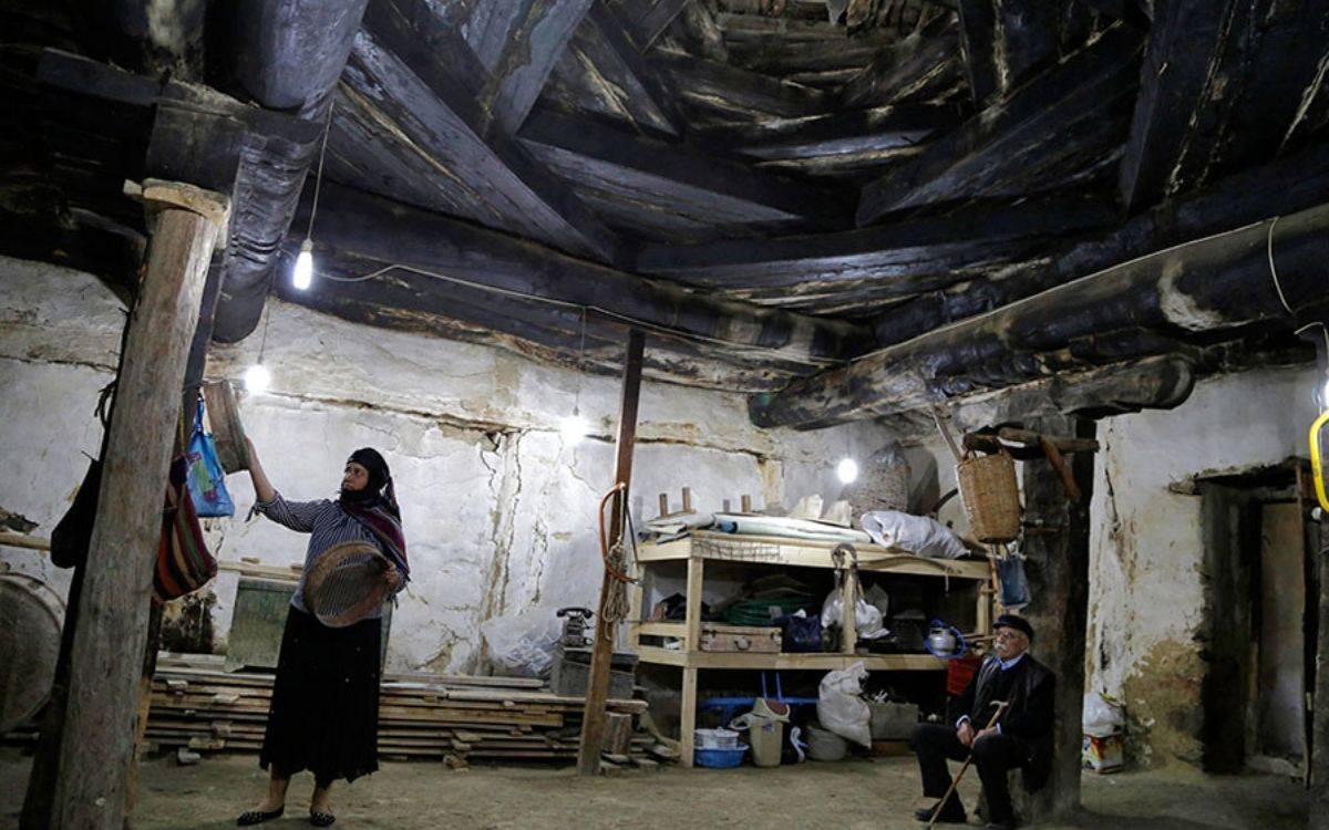 Fotoğrafta, geleneksel bir Anadolu evinin iç mekânı görülüyor. Odanın tavanı kalın, kararmış ahşap kütüklerle örülmüş; bu, yapının oldukça eski ve el emeğiyle inşa edildiğini gösteriyor. Duvarlar sıvalı ama yer yer çatlamış ve dökülmüş durumda. Zemini toprak olan bu alan, sade ama işlevsel bir yaşam alanı hissi veriyor.