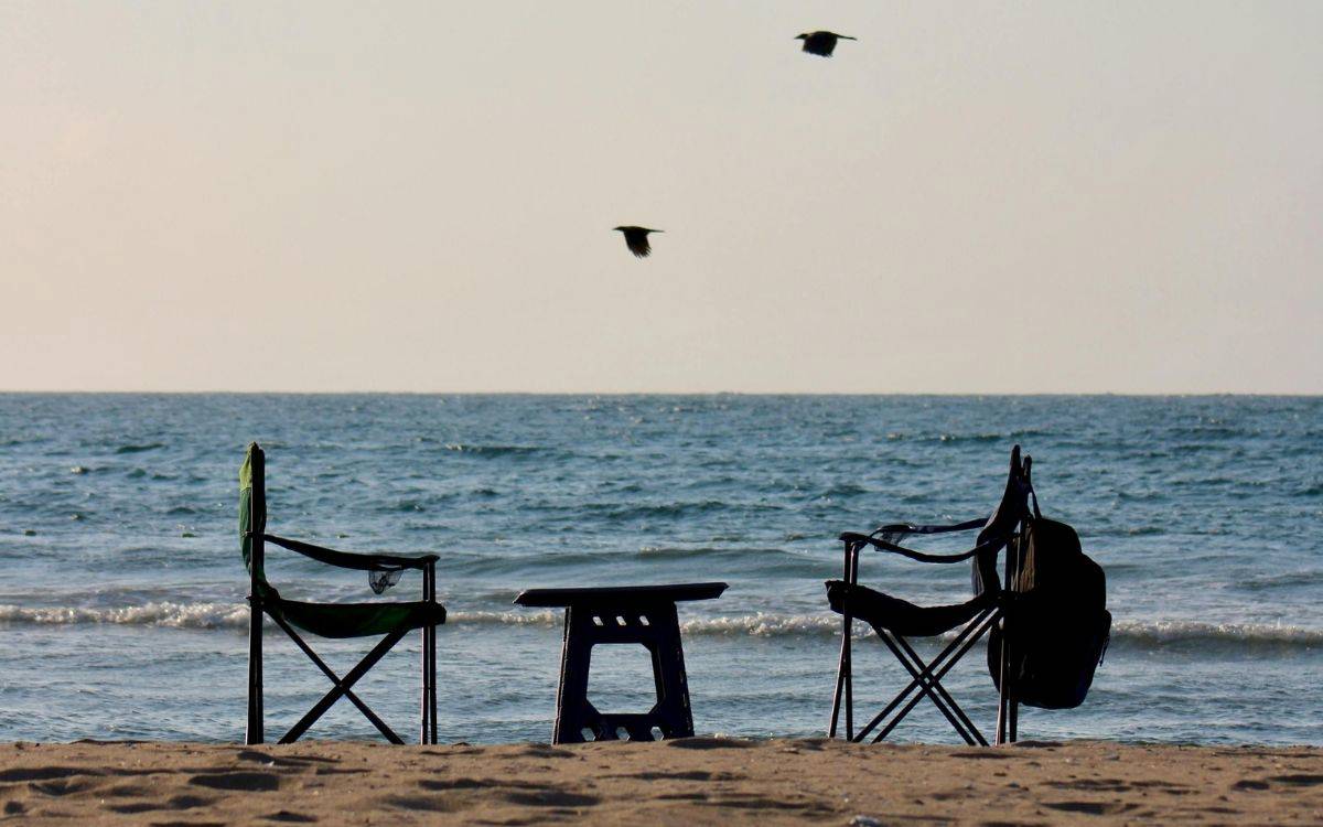 Fotoğrafta deniz kenarında, kumsal üzerinde yerleştirilmiş iki kamp sandalyesi ve aralarında duran küçük bir plastik masa görülüyor. Sandalyeler birbirine dönük şekilde konumlandırılmış; biri yeşil, diğeri ise siyah renkte. Sağdaki sandalyenin arkasına bir sırt çantası asılmış.