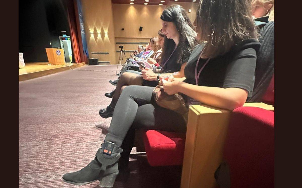 Görselde, bir etkinlik veya konferans salonunda oturan birkaç kadın var. Arka planda, bir sahne ve salonun düzeni gözüküyor. Ön planda ise birkaç kadın, rahat bir şekilde oturmuş ve cep telefonlarına bakıyorlar. Kadınlardan birinin ayağında, dikkat çekici şekilde bir elektronik kelepçe bulunuyor. Diğer katılımcılara kıyasla, bu detay, görseldeki en belirgin öge.