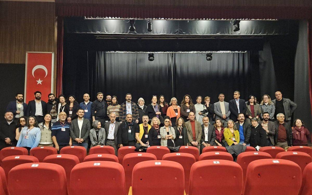 Fotoğrafta büyük bir grup insan, bir salonun sahne önünde toplu hâlde poz veriyor. Ön planda kırmızı renkli tiyatro/konferans koltukları var; insanların çoğu bu koltukların hemen arkasında ayakta duruyor, bazıları ise oturmuş durumda. Arka planda siyah perdeli bir sahne bulunuyor; tavanda sahne ışıkları görünüyor. Sol tarafta büyük bir Türk bayrağı asılı. Grup; kadınlar ve erkeklerden oluşan, yaşları farklı birçok kişiyi içeriyor. Çoğunun boynunda etkinlik kartı ya da yaka kartı bulunuyor; bu da fotoğrafın bir etkinlik, toplantı ya da konferans sonrası toplu hatıra fotoğrafı olduğunu düşündürüyor.