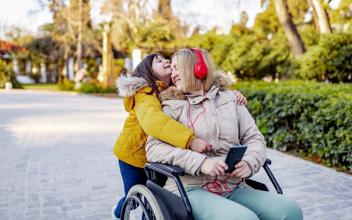 Fotoğrafta, güneşli bir günde parkta geçen sıcak ve neşeli bir an görülüyor. Kadrajın merkezinde bir kadın ve bir çocuk var. Kadın bir tekerlekli sandalyede oturuyor; açık renk bir mont giymiş, kulağında parlak kırmızı kulaklıklar var ve elinde bir akıllı telefon tutuyor. Yüzünde büyük, içten bir gülümseme var.