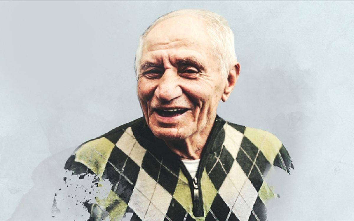 Fotoğrafta, arka planı açık gri tonlarda, sade bir zemin olan bir portre görüyoruz. Kadrajda yaşlı bir erkek var. Saçları kısa, beyaz ve seyrek; alnında ve yüzünde yaşlılığa bağlı belirgin çizgiler, kırışıklıklar var. Yüzünde kocaman, içten bir gülümseme görülüyor; ağzı hafifçe açık, gözleri gülmekten kısılmış, çok neşeli ve sıcak bir ifade taşıyor. Üzerinde baklava desenli bir kazak var; kazakta krem, siyah ve yeşil tonlarında geniş romb desenler bulunuyor. Fotoğrafın alt kısmında, sanki fırça darbeleriyle silikleşmiş ya da boyanmış gibi hafif bir efekt var; bu da portreyi daha yumuşak, neredeyse resimsi bir görünüşe kavuşturuyor. Genel olarak fotoğraf, sevecen, hayat dolu bir yaşlı insan hissi veriyor.