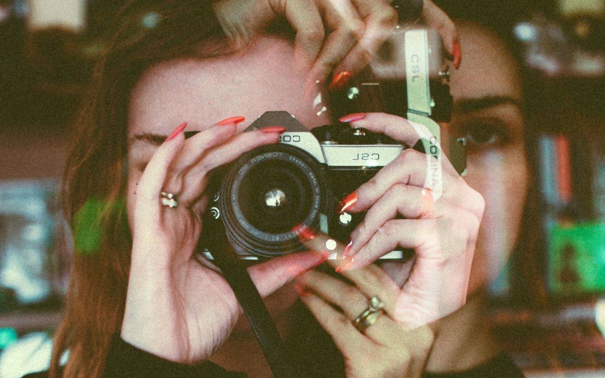 Fotoğrafta, kadraja çok yakın plan giren bir kişi görüyoruz; yüzünün büyük kısmı elindeki fotoğraf makinesiyle kapanmış. Eski tip, analog görünümlü, metal gövdeli bir fotoğraf makinesi tutuyor. Objektif tam ortada, izleyiciye doğru bakıyor; sanki o kişiyi değil, o kişi bizi çekiyormuş gibi.  Kişinin tırnakları uzun ve parlak kırmızı ojeli, birkaç parmağında yüzükler var. Fotoğrafta belirgin bir çift pozlama/üst üste binmiş görüntü efekti var: Eller, makine ve yüz iki kez görünüyormuş gibi; bir siluet diğerinin içine karışmış. Sağ tarafta, yarım beliren ikinci bir yüz ve göz dikkat çekiyor.