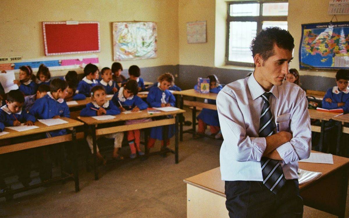 Sınıf ortamında çekilmiş bir fotoğraf bu. Önde, sağ tarafta genç bir erkek öğretmen ayakta duruyor. Kollarını göğsünde kavuşturmuş, düşünceli ya da biraz yorgun bir ifadeye sahip. Üzerinde açık renk gömlek, kravat ve kumaş pantolon var. Öğretmenin önünde bir öğretmen masası duruyor, üzerinde kâğıtlar görünüyor.