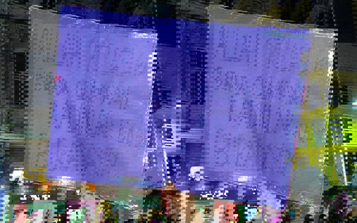 Görüntüde, bir kadın eylemde elinde mor renkli bir pankart tutuyor. Pankartın üzerinde, büyük harflerle yazılmış şu ifade yer alıyor: "ALLAH RIZASI İÇİN BURADA OLAMAYAN KADINLAR İÇİN DE BURADAYIM." Bu mesaj, kadının, toplumsal cinsiyet eşitliği ve kadın hakları için mücadeleye verdiği desteği simgeliyor.