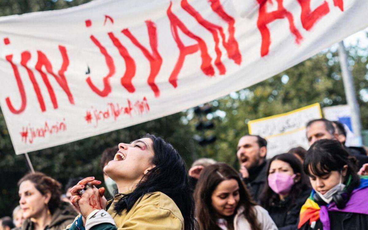Fotoğrafta bir protesto sahnesi görülüyor. Ön planda, duygusal olarak oldukça yoğun bir an yaşayan bir kadın var; başı hafifçe geriye dönük, ağzı açık şekilde bağırıyor ya da slogan atıyor. Elleri kenetlenmiş, yüzündeki ifade güçlü bir öfke, acı ve kararlılık karışımı hissettiriyor. Üzerinde açık renk bir mont bulunuyor.