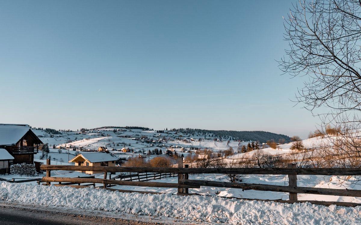 Karlarla kaplı yayla veya kırsal bir bölge uzanıyor. Yamaçlar yumuşak, tepeler hafifçe yükselip alçalıyor. Ahşap evler ve kulübeler dağınık hâlde manzaranın içinde yer alıyor; çatılarında kalın kar birikmiş. Ön planda tahta bir çit uzanıyor ve yol kenarında kürenmiş kar yığınları bulunuyor. Arka planda ise çam ormanları ve küçük yerleşim alanları görülüyor. Gökyüzü açık, pembe-mavi tonlu bir gün batımı veya gün doğumu ışığı manzaraya yumuşak bir sıcaklık katıyor.