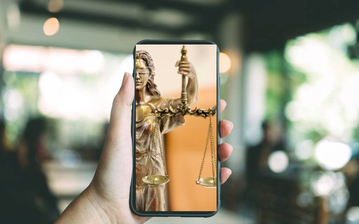 Görüntüde, bir kişinin elinde tuttuğu akıllı telefonun ekranı yakından görülüyor. Telefonun ekranında, adaletin simgesi olan Themis heykelinin bir görüntüsü yer alıyor: Gözleri bağlı, bir elinde terazi, diğer elinde kılıç tutan klasik adalet figürü. Heykelin özellikle terazi kısmı ve yüzünün bir bölümü ekranda net şekilde görünmekte.