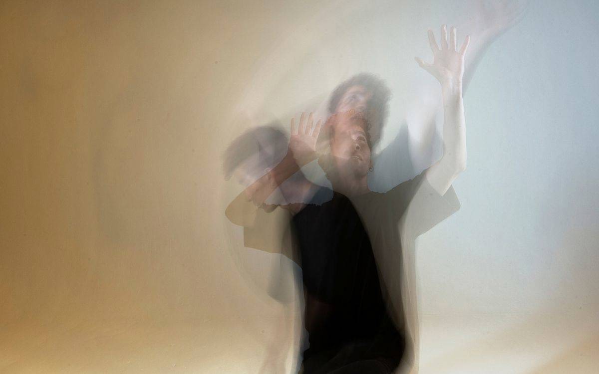 Görselde açık renkli, boş bir duvarın önünde duran bir kişi yer alıyor. Fotoğraf bilinçli olarak bulanık ve çoklu pozlama efektiyle çekilmiş; bu nedenle aynı kişinin üst üste binmiş birkaç silueti görülüyor. Kişi siyah bir tişört giymiş. Kollarını yukarı doğru kaldırmış; elleri açık ve parmakları gerilmiş durumda. Hareket hâlindeymiş gibi görünüyor. Başının ve kollarının farklı yönlere kaymış, yarı saydam görüntüleri üst üste geçmiş; bu da güçlü bir hareket ve dağılma hissi yaratıyor.
