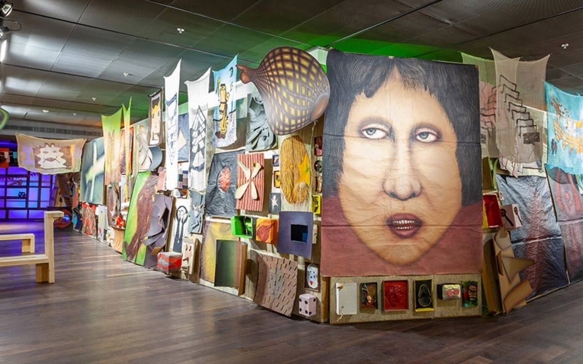 Fotoğraf, kapalı bir sergi alanında kurulmuş yoğun ve katmanlı bir enstalasyonu gösteriyor. Uzun bir duvar boyunca birbirine eklenmiş çok sayıda resim, kumaş parçası, küçük obje ve üç boyutlu çalışmalar yerleştirilmiş. Alan, bir galeri ya da müze ortamını andırıyor; tavan aydınlatmalarıyla aydınlatılmış, zemini düz ve geniş bir yürüyüş alanı bırakılmış.