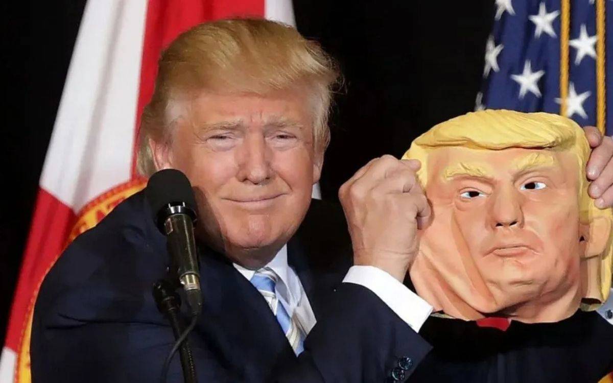 Fotoğrafta Donald Trump bir kürsüde konuşma yaparken görülüyor. Önünde bir mikrofon var ve arka planda ABD bayrağı dikkat çekiyor. Trump koyu renk takım elbise ve açık renk kravat giymiş.